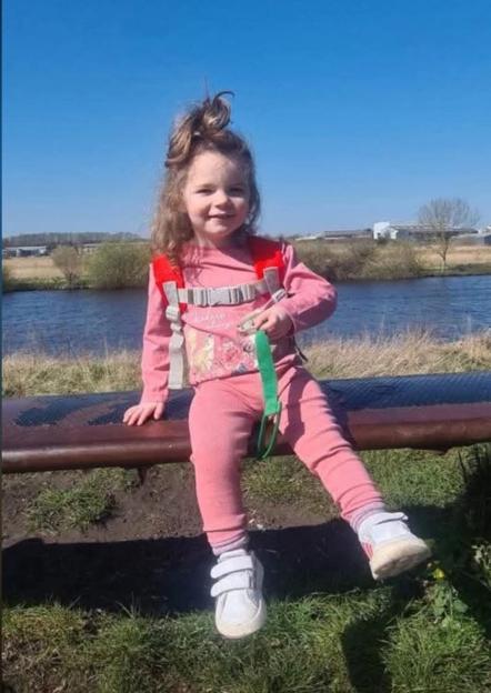 Isabelle Rose Welsh, a young girl with curly hair and a pink outfit, sitting on a bench with a body of water behind her.