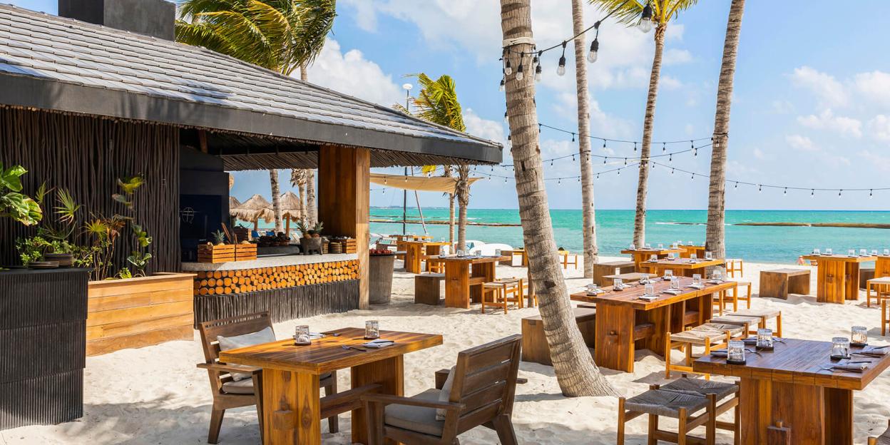 Fairmont Mayakoba luxury hotel's outdoor restaurant with wooden tables and chairs on a sandy beach next to turquoise water and palm trees.