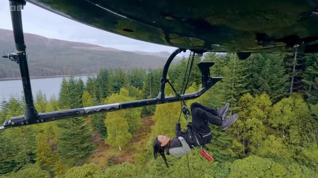 Maya Jama hanging from a helicopter above a forest and lake.