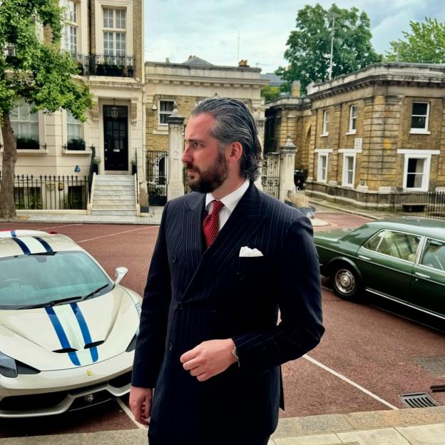 Ricky Lawrence in a pinstriped suit, standing outdoors with a cream sports car and a dark green vintage car in the background.