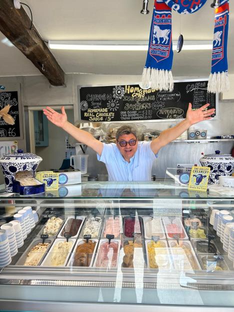 Ugur Vata, also known as the "Willy Wonka of ice cream", with his arms outstretched behind a display case of various ice cream and sorbet flavors in his shop, The Gallery Creamery and Deli.