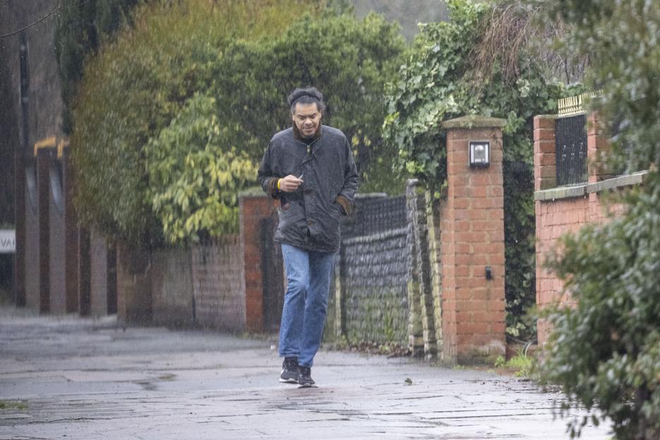 Daniel Bannister, wearing a dark jacket and jeans, walks down a paved driveway on a rainy day.