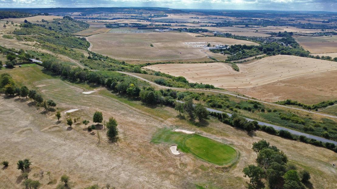 Aerial view of the Dunstable Downs Golf Club course with a vivid green putting green contrasting with drought-affected brown grass, next to a road and fields, with a glider visible in the sky.