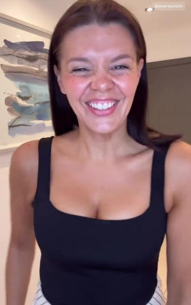 A woman with brown hair and a black top is laughing in front of a painting.