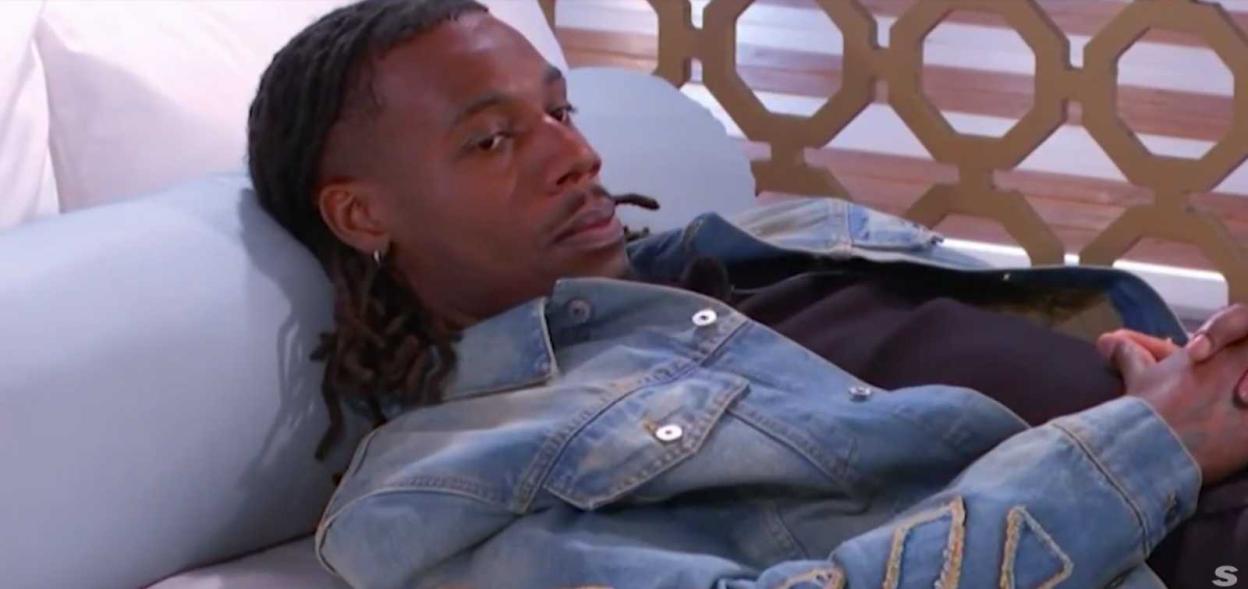 A man with dreadlocks lies on a bed wearing a denim jacket, looking off to the side with a pensive expression.