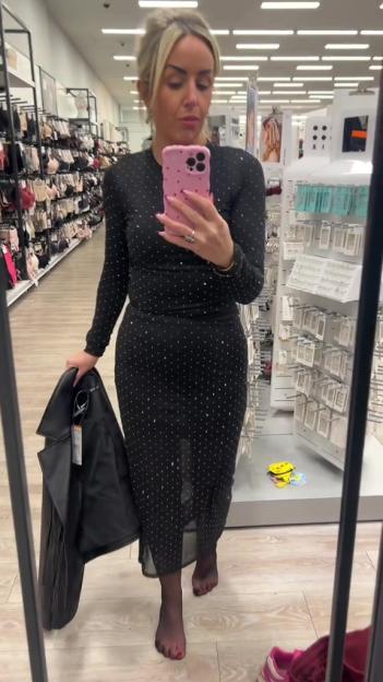 Woman in a black dress with rhinestones holding a black leather jacket, looking into a mirror in a store.