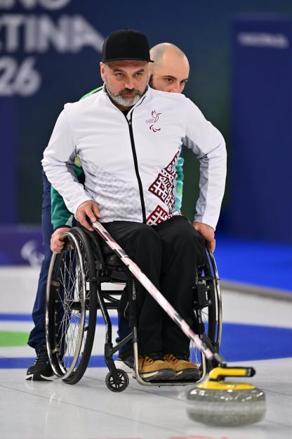 A male wheelchair curler aims a shot while a teammate stands behind him for support during a Paralympic game.