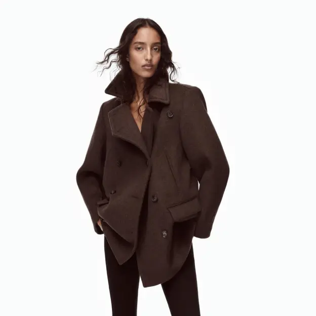 A woman wearing a brown double-breasted coat and dark pants against a plain white background.