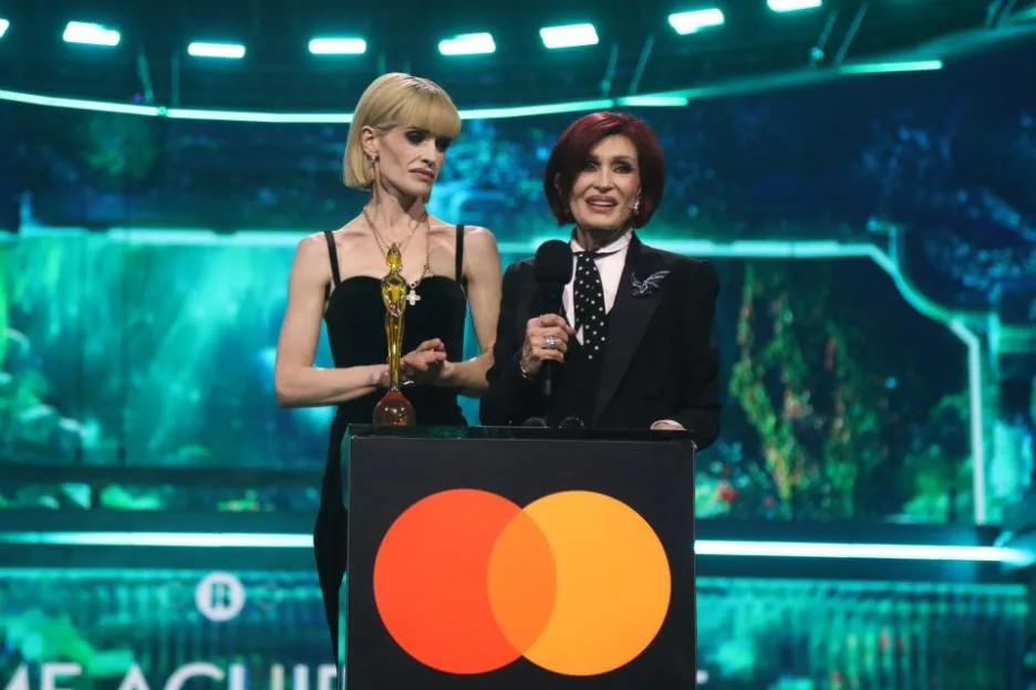 Two women on stage at the BRIT Awards, with one holding a microphone behind a podium bearing the Mastercard logo.