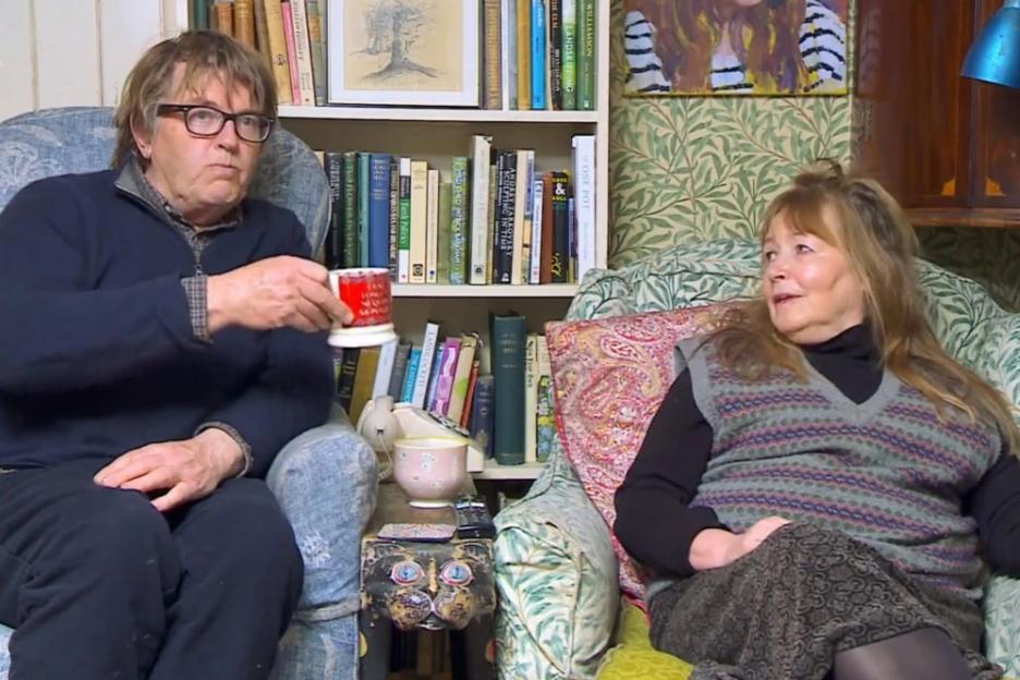 Mary and Giles sitting in armchairs in a living room filled with books.