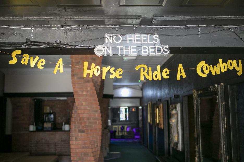 A neon sign reading "NO HEELS ON THE BEDS" and a yellow banner reading "Save A Horse Ride A Cowboy" hanging in a doorway.