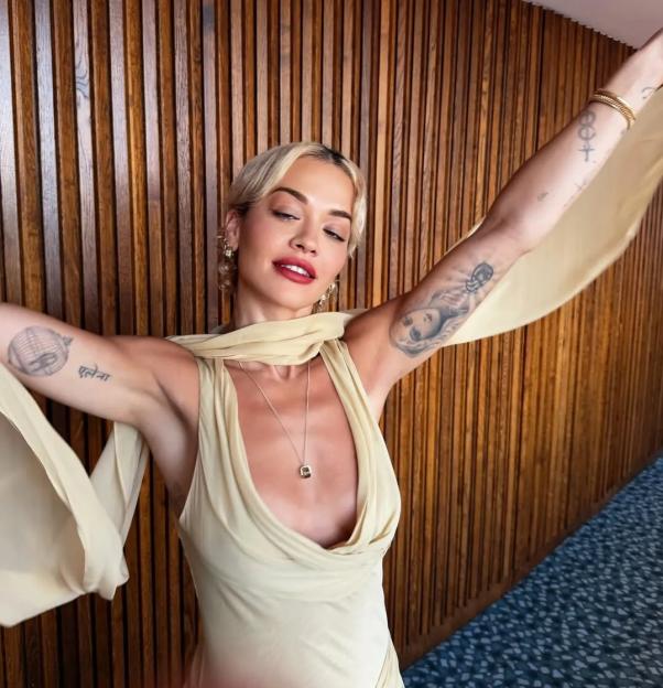 Rita Ora in a plunging beige top with arms outstretched.