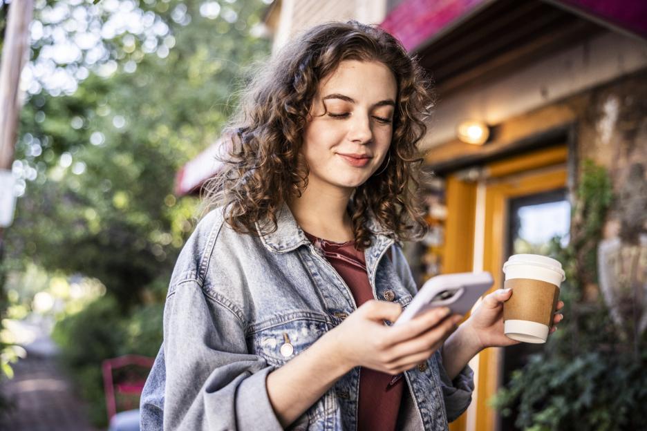 A young woman with curly brown hair, wearing a denim jacket and a red top, stands outdoors, holding a phone in her left hand and a coffee cup in her right.