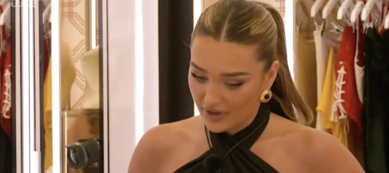 Belle Hassan talking with a serious expression, wearing a black top and gold earrings, with clothes hanging in the background.