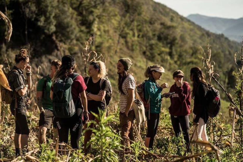 A group of hikers with backpacks stand together in a field with green hills in the background.