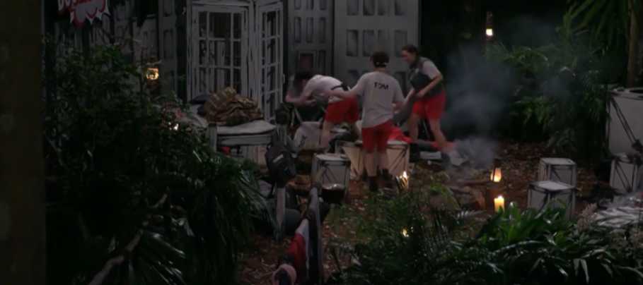 Shona and Aitch from IAC in red shorts and grey t-shirts engaged in an activity in a jungle camp, with smoke and candlelight.