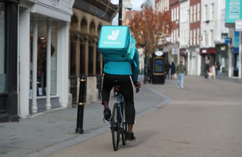 A Deliveroo delivery person cycling away from the camera on a city street.