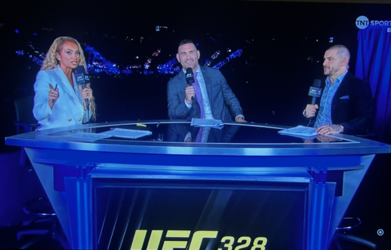 Three commentators, one woman and two men, seated at a blue desk with "UFC 328" on the front.