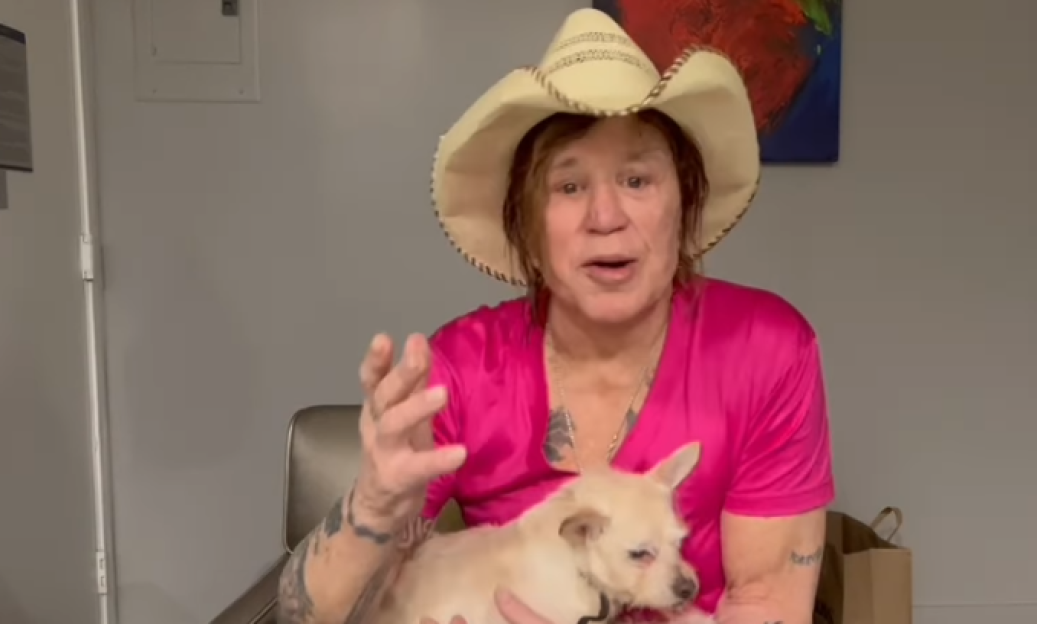 Mickey Rourke in a cowboy hat and pink shirt holding a small dog.