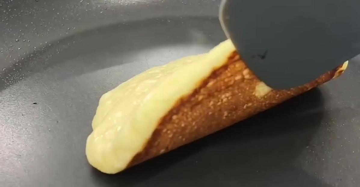 A pancake cooking in a frying pan being lifted by a spatula to reveal a golden-brown underside.