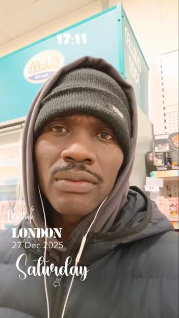 A man, Olabode Shoniregun, wearing a black beanie and hooded jacket with earbuds in, looks directly at the camera in a store in London.