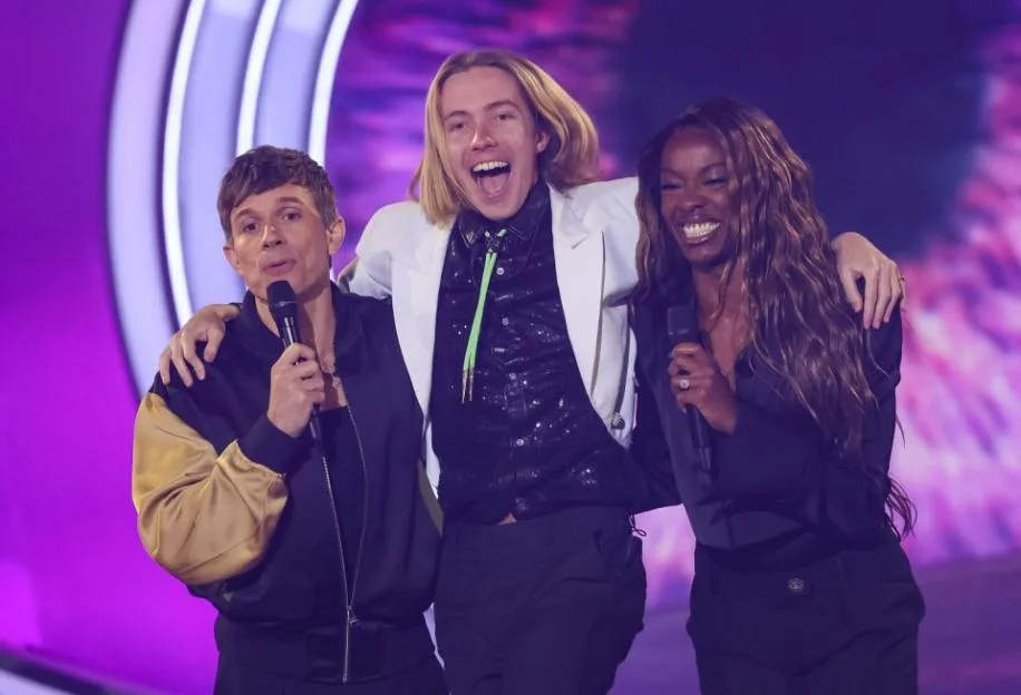 Three people smiling and holding microphones on stage, with the person in the middle, who has long blonde hair, being lifted up by the other two.