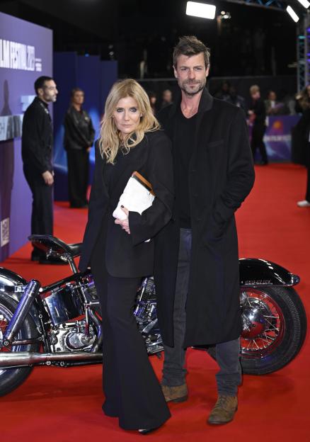 An older blonde woman and a younger man in black outfits stand next to a shiny black motorcycle on a red carpet.