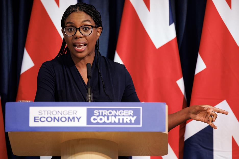 Kemi Badenoch delivering a speech on the UK economy, standing at a podium with "STRONGER ECONOMY STRONGER COUNTRY" written on it, with Union Jack flags in the background.