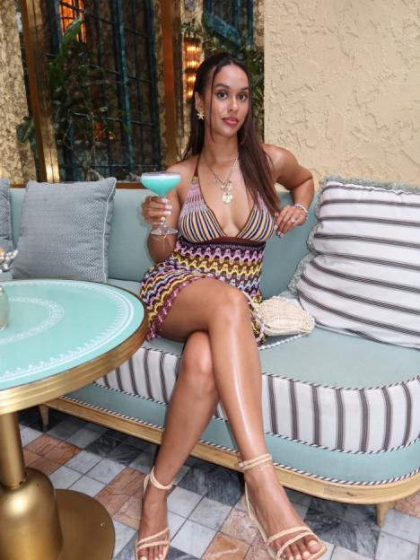 Woman in a colorful knit dress holding a blue cocktail while sitting on a teal bench.