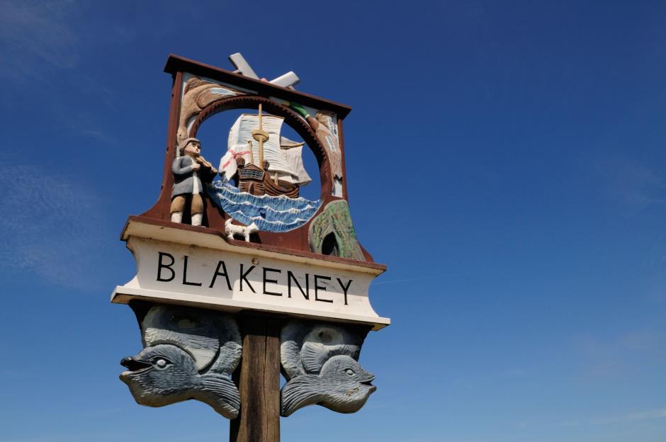 Carved wooden Blakeney Village sign in Norfolk, England, UK, featuring a man playing a violin, a sailboat, a dog, and two fish.