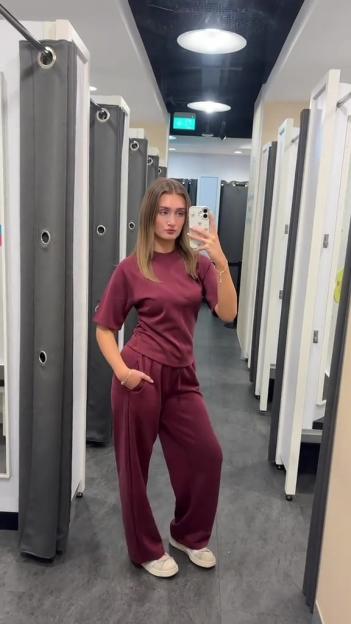 A young woman poses in a fitting room wearing a maroon short-sleeved top and matching wide-leg pants.
