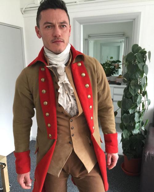 Luke Evans in a historical costume with a tan coat, red lapels, and a ruffled white shirt.