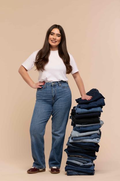 A woman in a white t-shirt and blue jeans smiles while leaning on a stack of folded jeans.