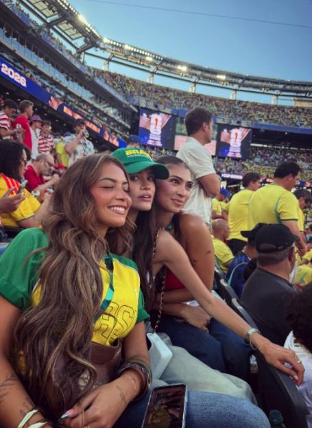 Three women in a stadium, with two wearing Brazil fan gear, smiling at a sporting event.