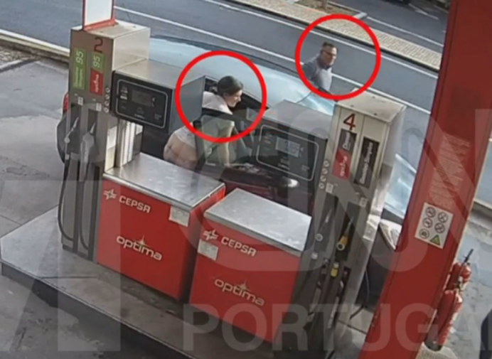 A woman with her pants down is crouched behind a gas pump, while a man is seen in the background.