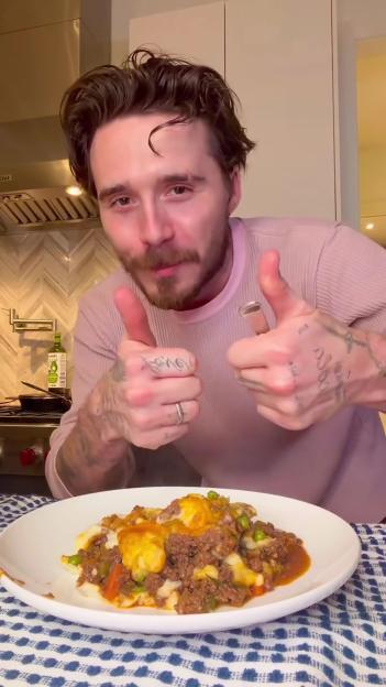 Brooklyn Beckham in a kitchen with a plate of shepherd's pie, giving two thumbs up.