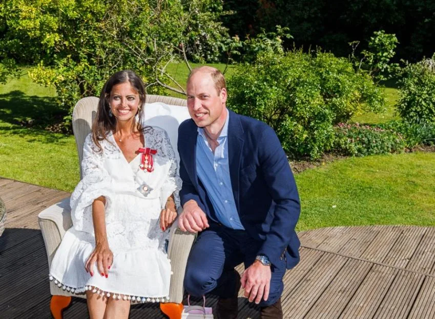 Deborah James wearing her Damehood medal seated in a chair next to Prince William outdoors.