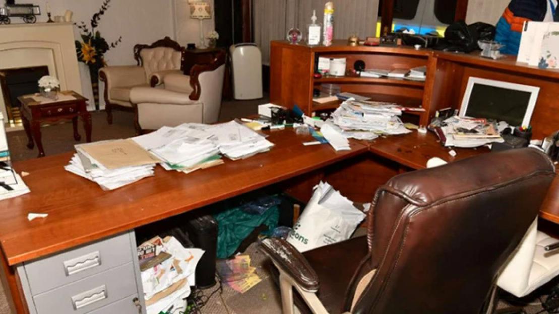 A messy office desk with stacks of papers and a computer, in front of a living room area with a fireplace and ornate chairs.