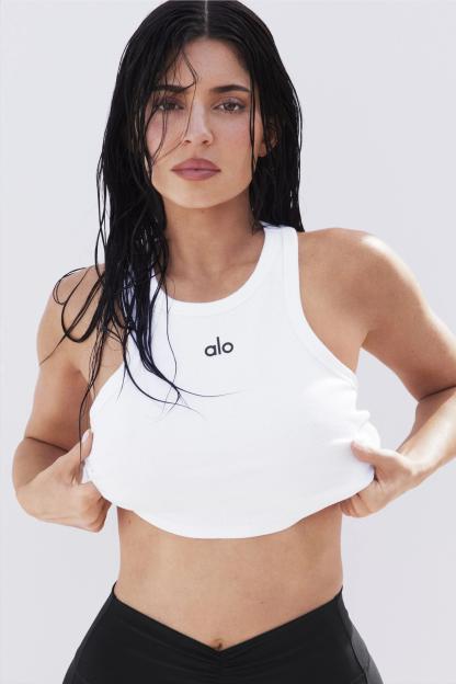 Kylie Jenner in an Alo Yoga campaign, wearing a white ribbed tank top and black bottoms, pulling up the bottom of her top with both hands.