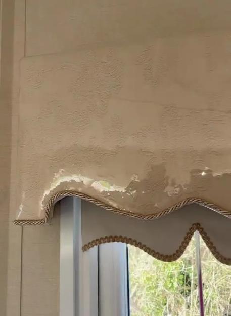 An ornamental pelmet, protected by plastic wrap, above a window with a green outdoor view.