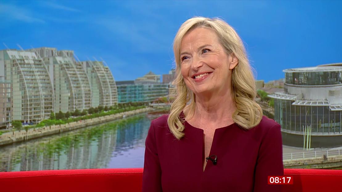 A blonde woman in a burgundy top speaks in front of a backdrop of a city with a river.