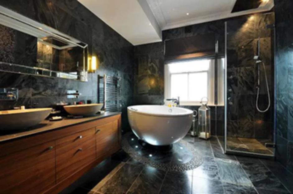 An interior view of a modern bathroom featuring dark slate tile walls and floors, a freestanding white bathtub, a double wooden vanity unit with vessel sinks, and a glass-enclosed shower.