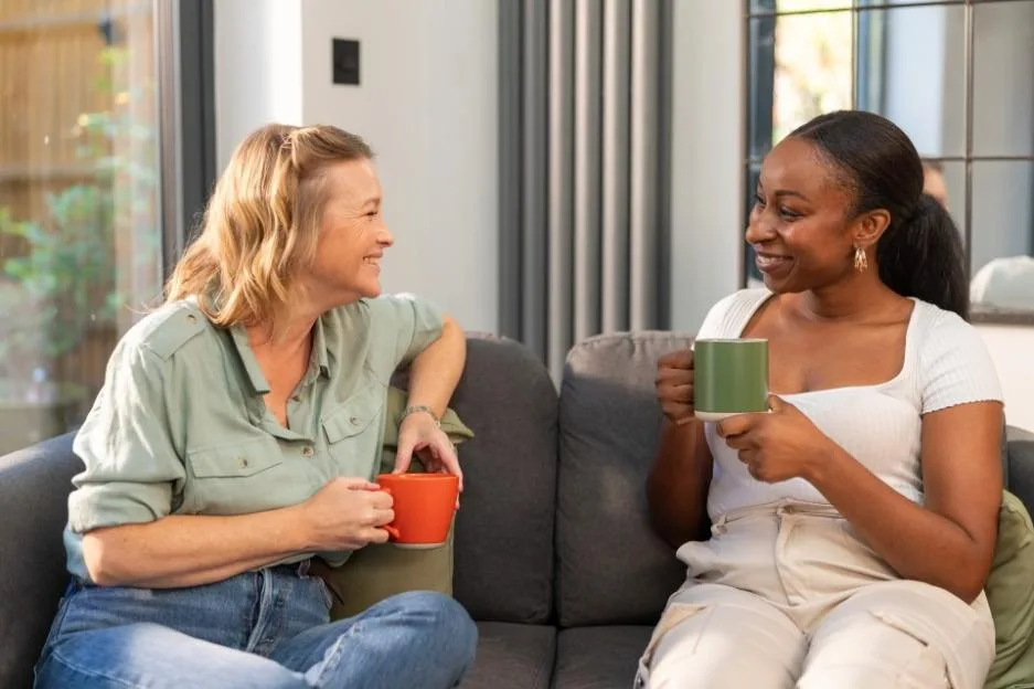 Two women sitting on a sofa, chatting and holding mugs.