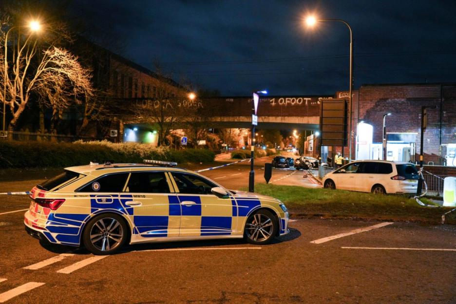 An evening street scene in Birmingham with a checkered police car in the foreground, and a cordoned-off crash site under a bridge in the background.