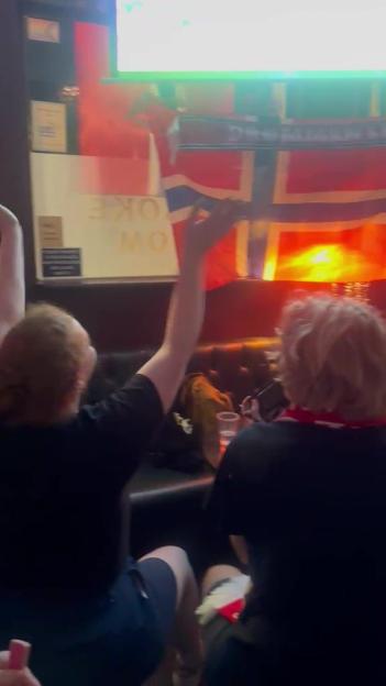 People in a dimly lit bar celebrating in front of a screen and Norwegian flags.