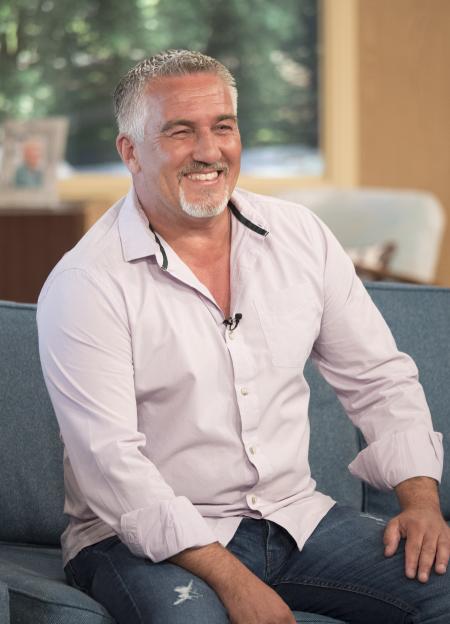 Paul Hollywood smiling and looking to the left, wearing a light purple button-up shirt, seated on a blue couch.