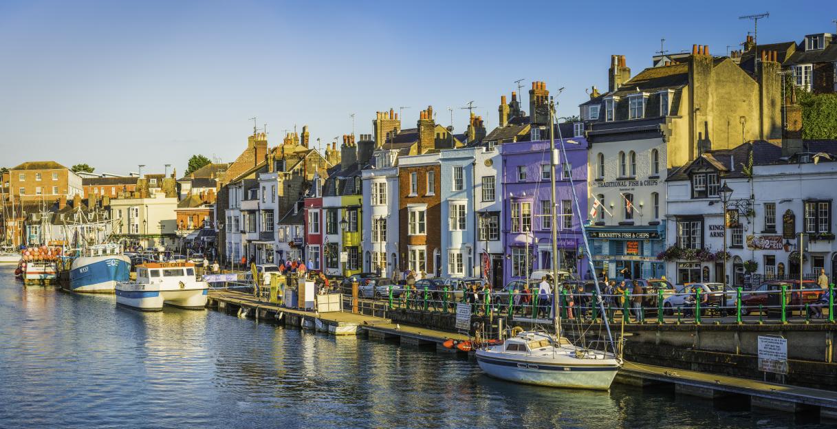 Colorful cottages, fishing boats, and yachts in the harbor of Weymouth, Dorset, with people enjoying the warm summer evening outside waterfront pubs and restaurants.
