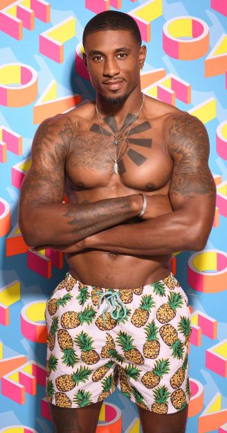 a shirtless man in pineapple swim trunks stands with his arms crossed