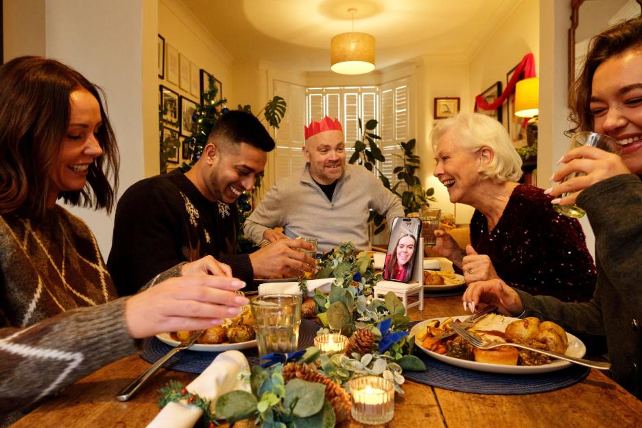 A family laughing at a table during a Christmas meal, with one person joining via video call on a phone.