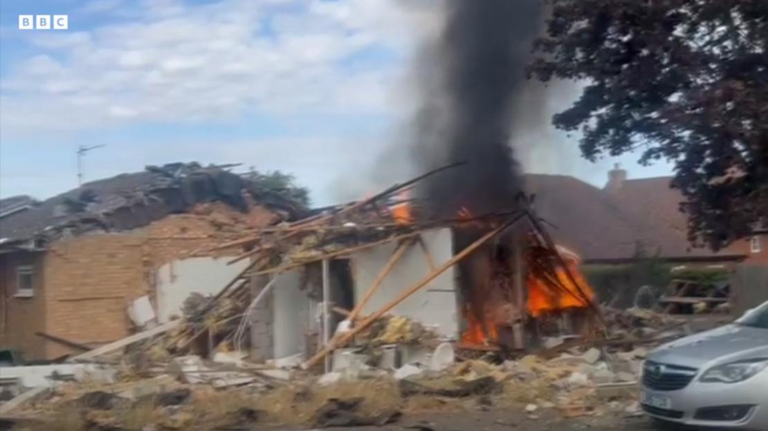 A destroyed bungalow with flames and black smoke billowing from its remains after a gas explosion.